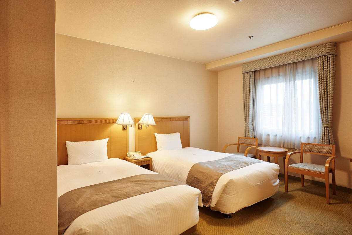 Quarto de hotel com duas camas, uma mesa redonda pequena, uma cadeira e uma janela com cortinas.