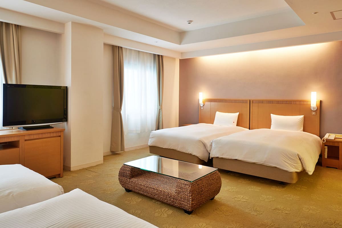 Quarto de hotel com duas camas, uma televisão sobre um armário de madeira, uma mesa de centro de vidro e uma luminária fixada na parede.