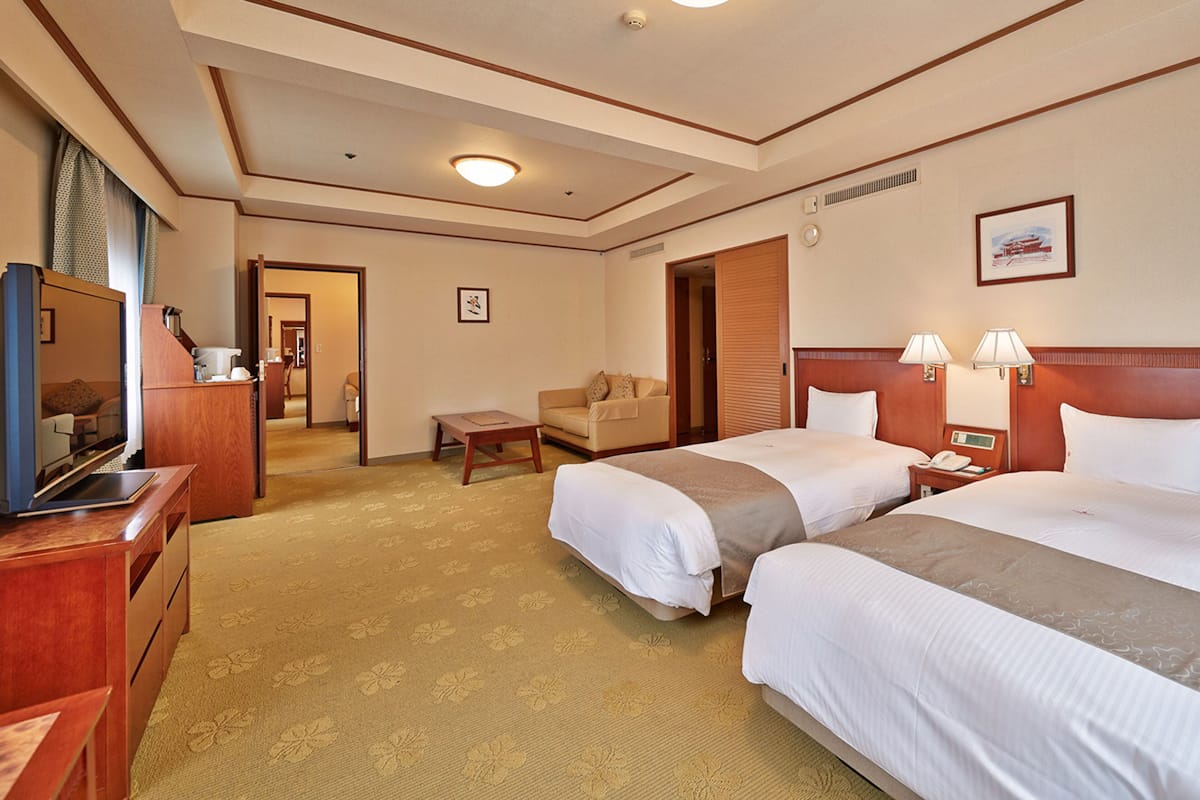 Quarto de hotel com duas camas, uma escrivaninha, televisão e um pequeno espaço de estar.