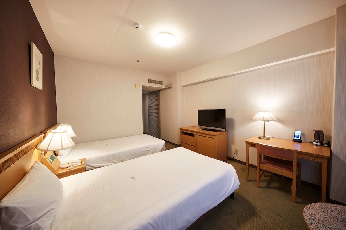 Quarto de hotel com duas camas de solteiro, uma escrivaninha com televisão, luminária e telefone.