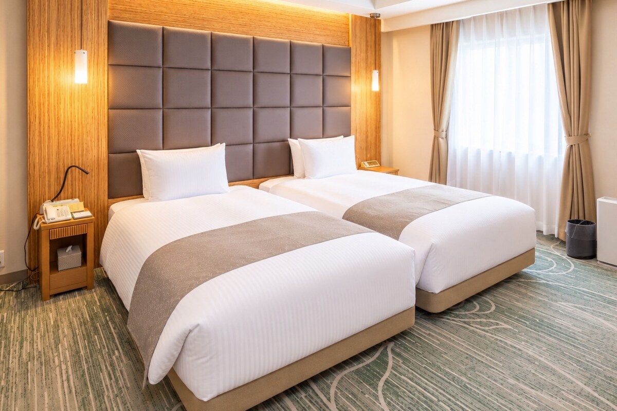 A hotel room with two beds, a wooden headboard, a bedside table with a lamp, and a window with curtains.