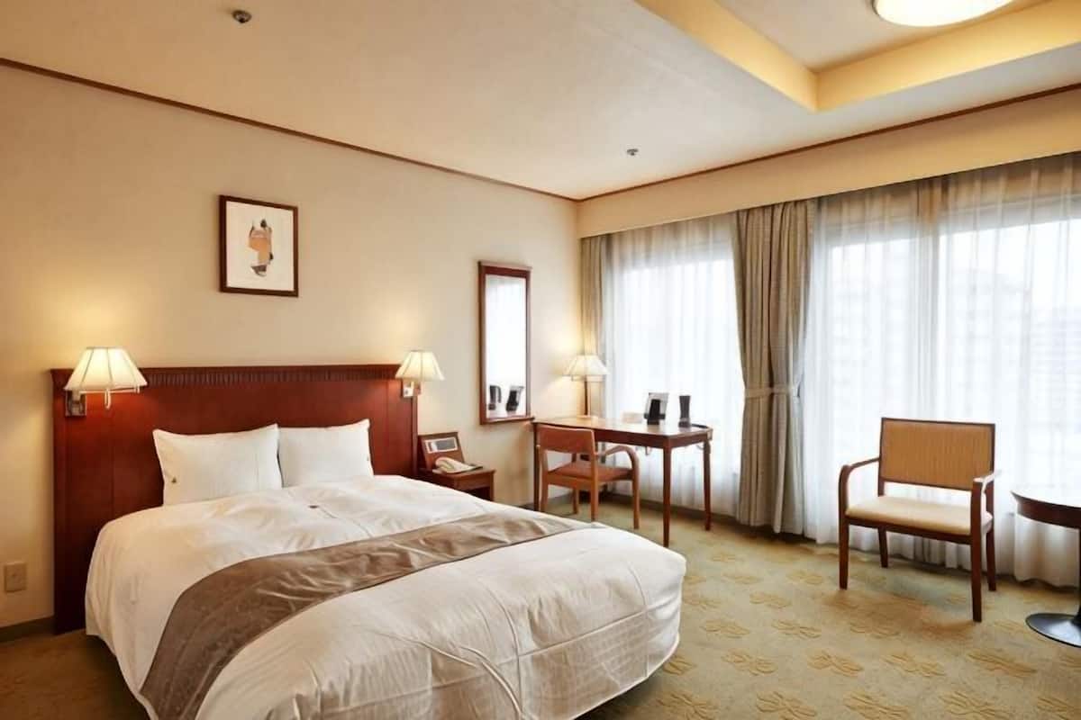 Quarto de hotel com uma cama grande, duas luminárias de cabeceira, uma escrivaninha, uma cadeira e uma janela ampla com cortinas.