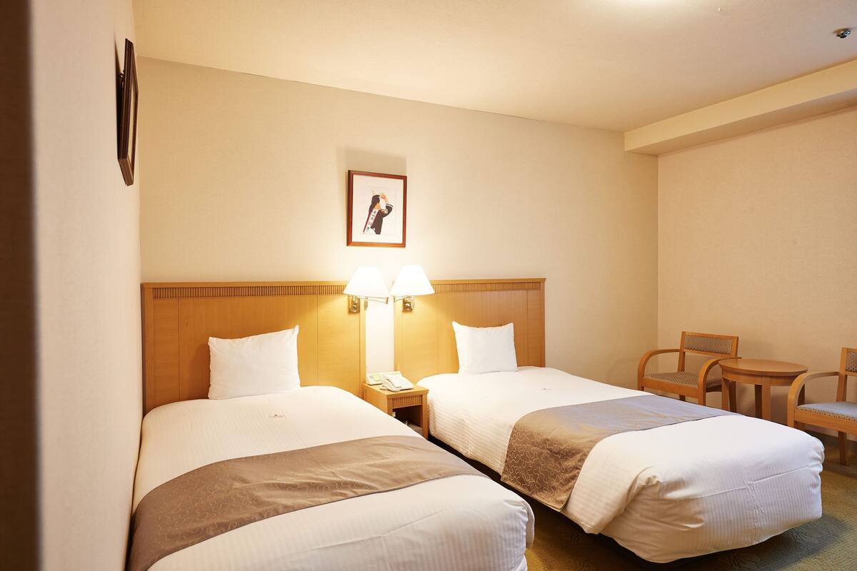 Quarto de hotel com duas camas, cabeceira de madeira, luminárias de cabeceira, uma mesinha e duas cadeiras.