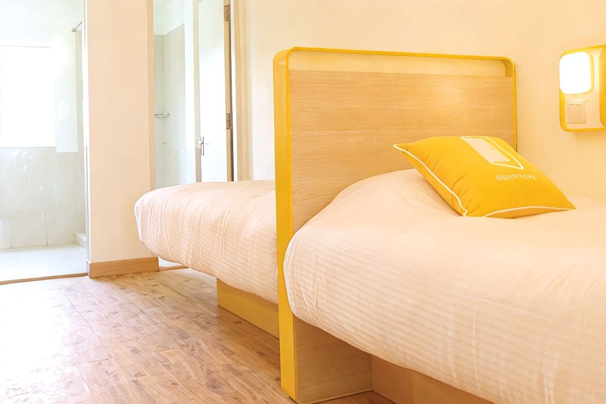 A single bed with a wooden frame and a yellow pillow, a bedside table, and a wall-mounted light fixture.