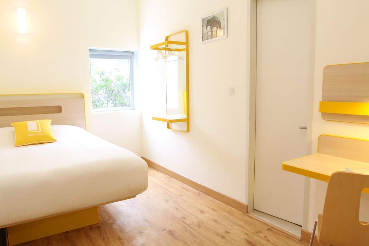 A bedroom with a bed, a bedside table, a chair, a window, a door, and a wall-mounted shelf.