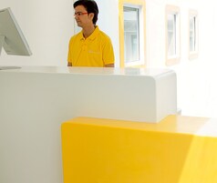 Un mostrador de recepción con un monitor de computadora, una persona con una camisa amarilla y un mostrador amarillo brillante.