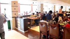 A busy restaurant with multiple dining tables and chairs, a central counter, and a large window in the background. - BKR Grand (Chennai)
