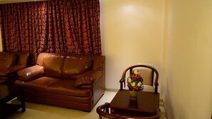 A living room with a brown leather sofa, a wooden dining table with a vase of flowers, and a patterned curtain. - BKR Grand (Chennai)