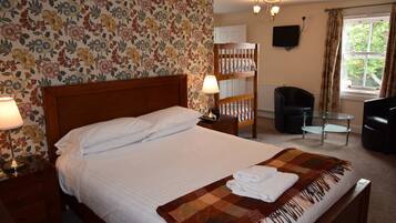 Habitación de hotel con cama, cabecera de madera, mesita de noche con lámpara, una pequeña mesa con una silla, televisor montado en la pared y ventana con cortinas.