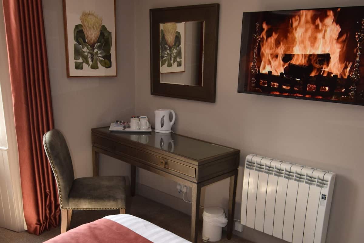 A hotel room with a bed, a desk, a chair, a radiator, and a wall-mounted TV displaying a fire.