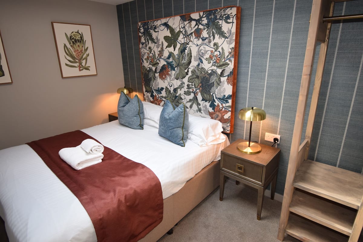 A neatly made bed with a red blanket, a nightstand with a lamp, and a floral tapestry on the wall.