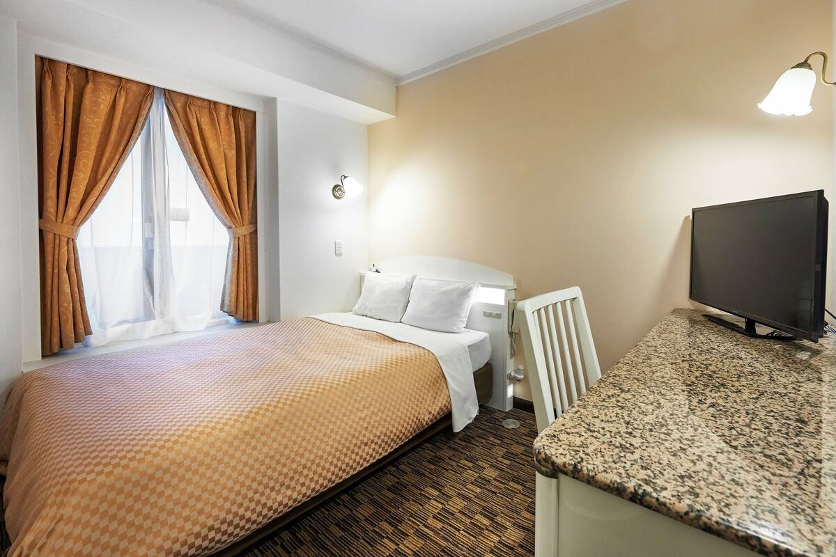 Una habitación de hotel con cama, escritorio con televisor, silla y ventana con cortinas.