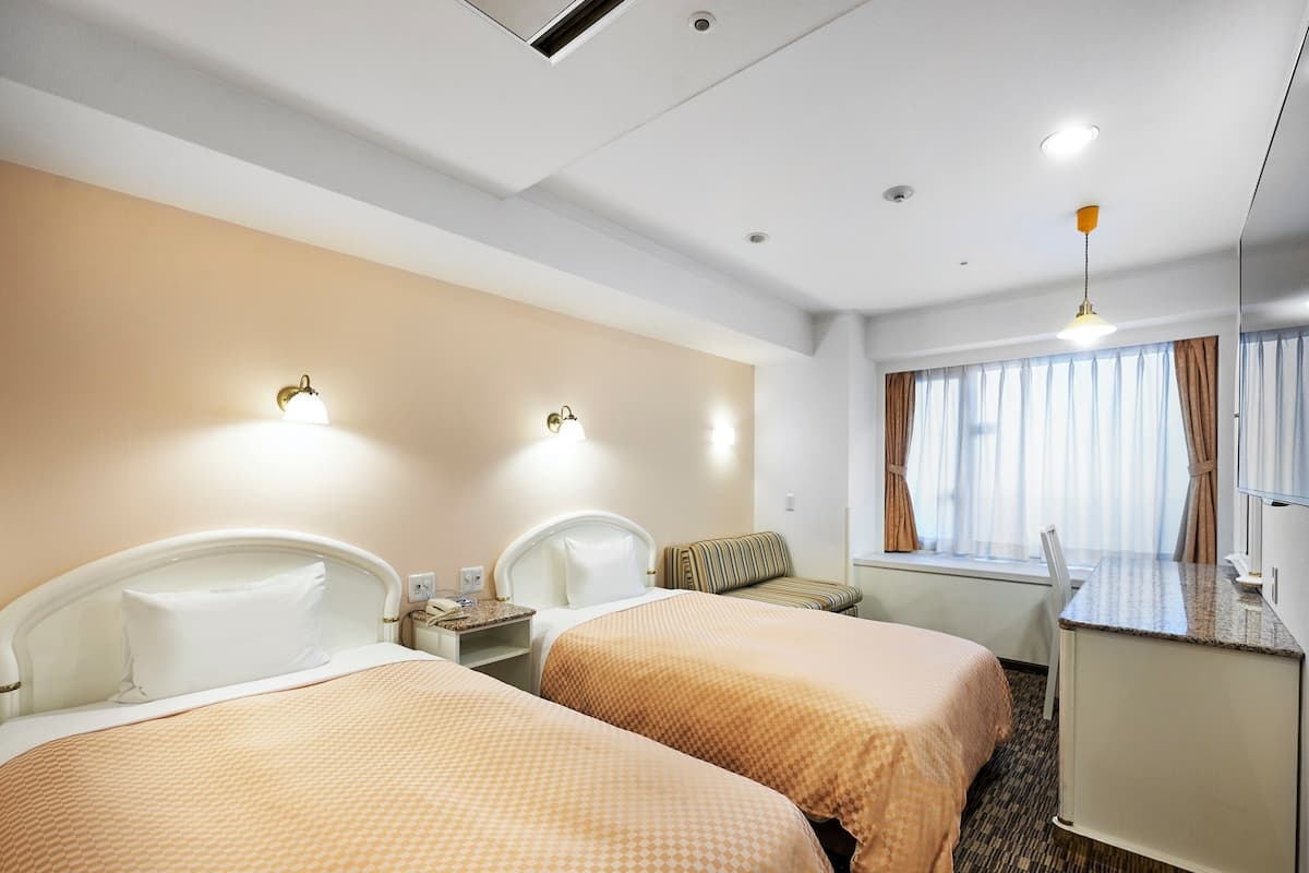 Habitación de hotel con dos camas, un escritorio pequeño, una silla y un ventanal con cortinas.