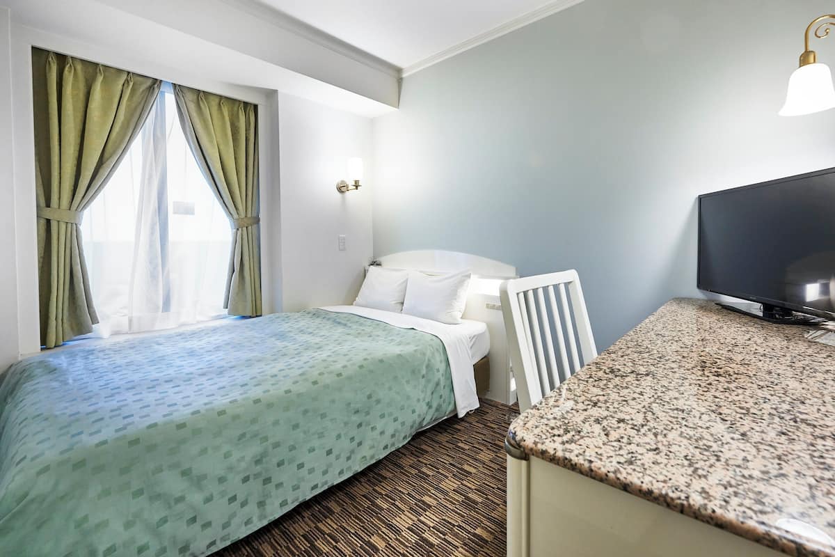 Una habitación de hotel con cama, escritorio, televisor y ventana con cortinas.