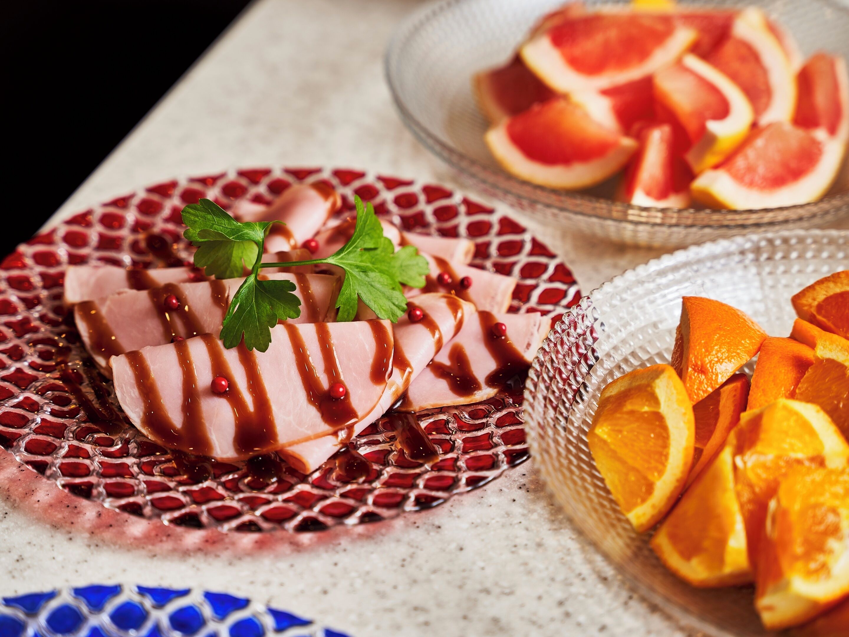 A plate of sliced meat with a garnish and sauce, a bowl of orange slices, and a decorative plate with a red and white pattern.