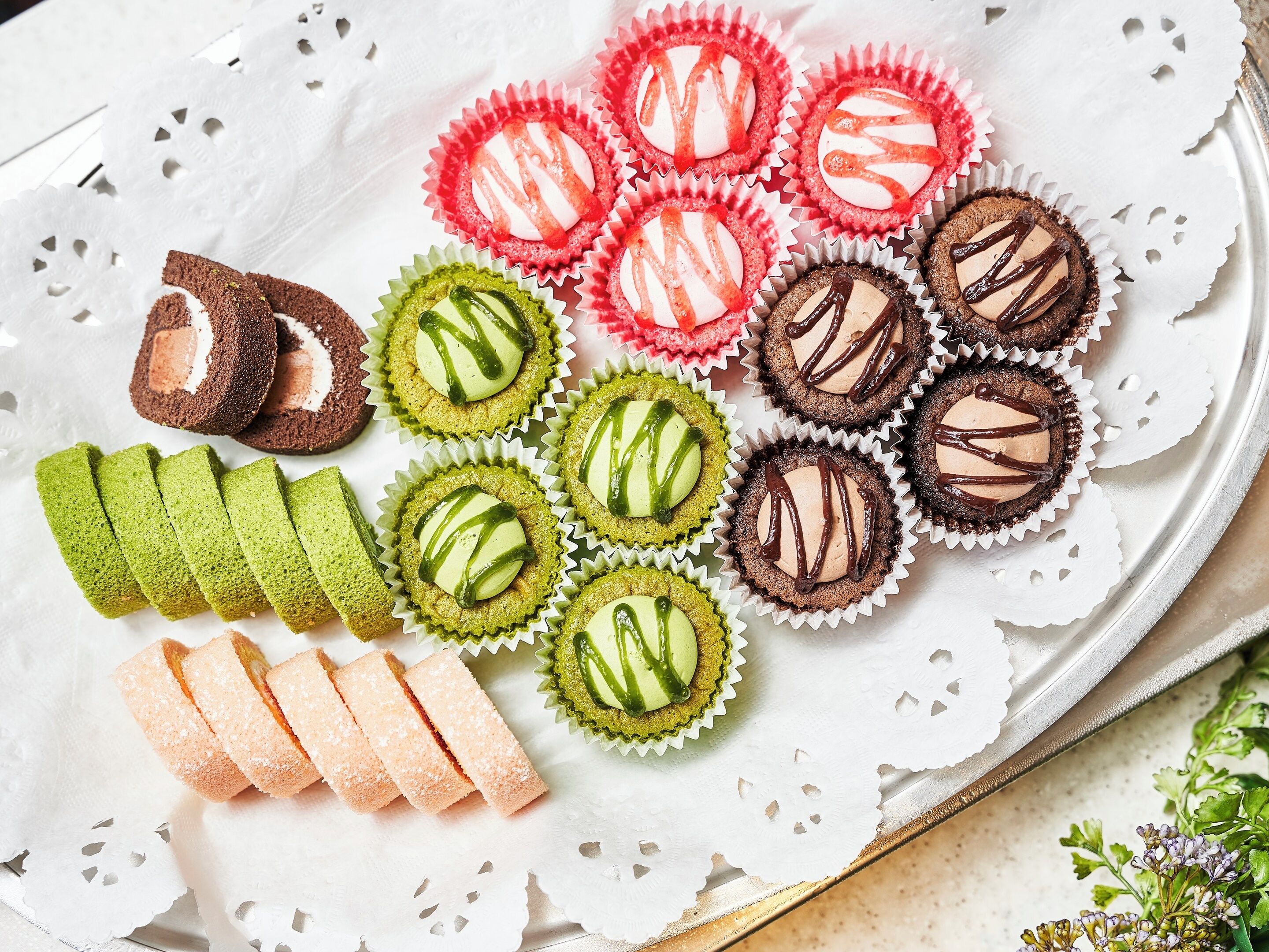 A tray of assorted pastries and cookies, including pink and green varieties, arranged on a white doily.
