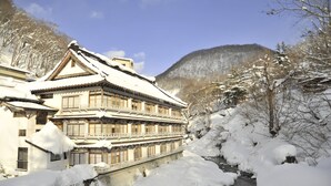 A traditional building with multiple windows and a snow-covered roof, situated by a river with snowbanks. - Takaragawa Onsen Osenkaku (Minakami)