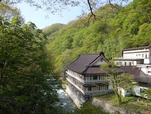 Een traditioneel Japans gebouw met houten constructie en stenen fundering, omgeven door weelderige begroeiing en een rivier.