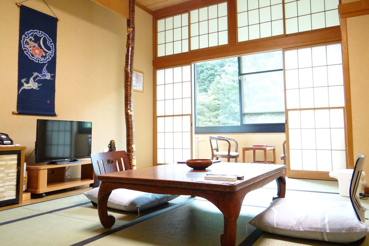 Una habitación tradicional japonesa con suelo de tatami, una mesa de madera baja, un televisor y un ventanal con vistas a zonas verdes.