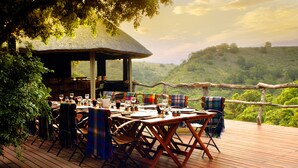 An outdoor dining area with wooden tables and chairs, a thatched-roof structure, and a scenic view of a lush green hillside. - Lalibela Game Reserve (Grahamstown)