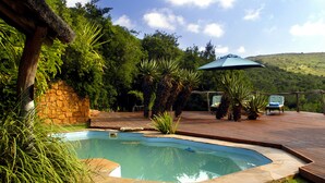 A swimming pool surrounded by lush greenery, a stone wall, and a wooden deck with chairs and an umbrella. - Lalibela Game Reserve (Grahamstown)