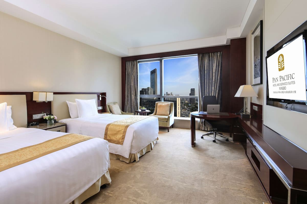 A hotel room with two beds, a desk, a chair, and a large window with a city view.