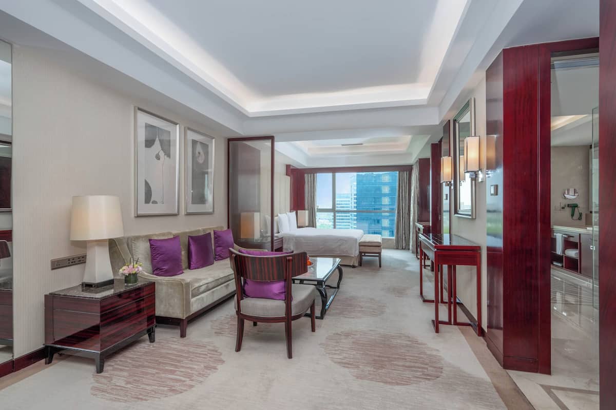 A hotel room with a sofa, armchair, bedside tables, a bed, a desk, and a large window offering a city view.