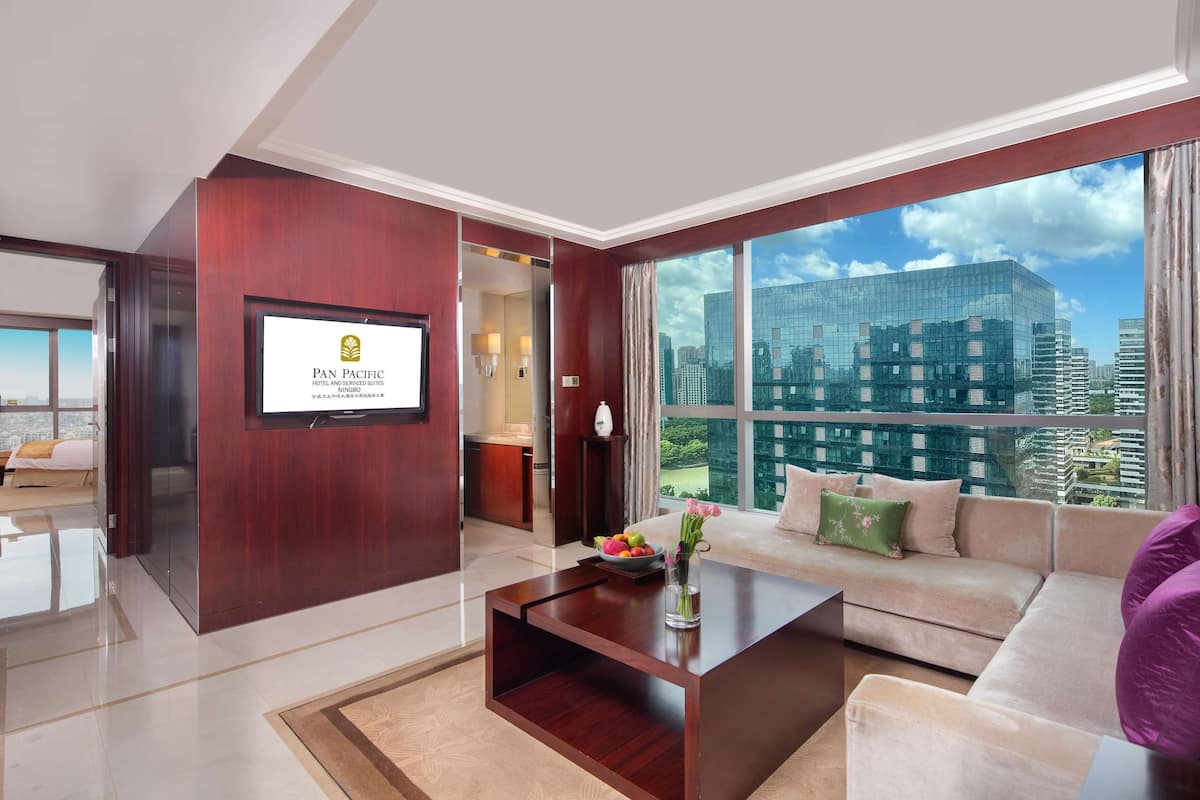 A modern hotel room with a large window offering a city view, a flat-screen TV mounted on the wall, and a comfortable seating area with a sofa and a coffee table.