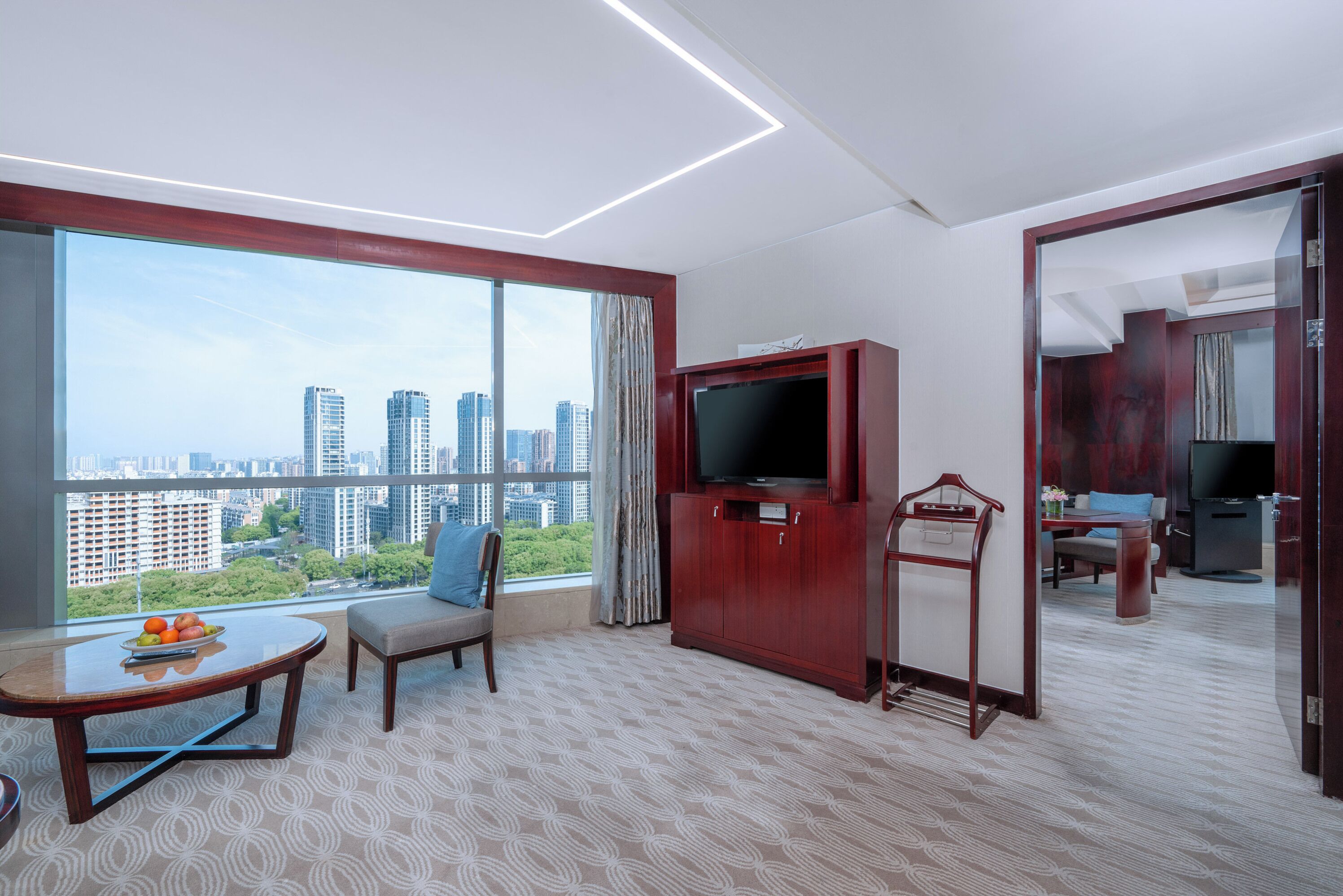 A hotel room with a large window offering a city view, a flat-screen TV, a dining table with fruit, and a seating area.