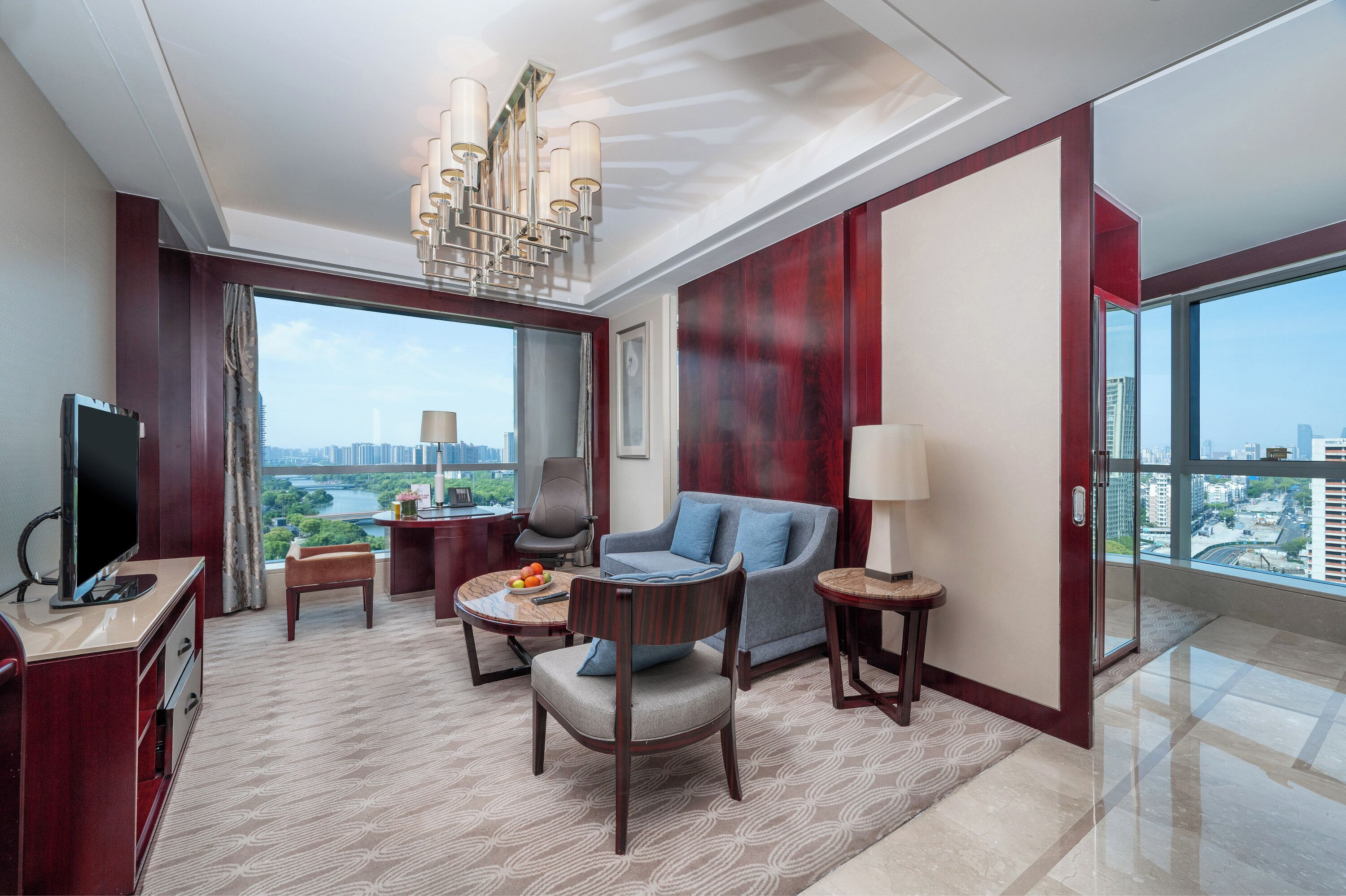 A modern hotel room with a large window, a flat-screen TV, a sofa, a chair, a small table, and a lamp.