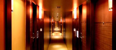 Pasillo de hotel con puertas de madera, iluminado por apliques, y una alfombra estampada.