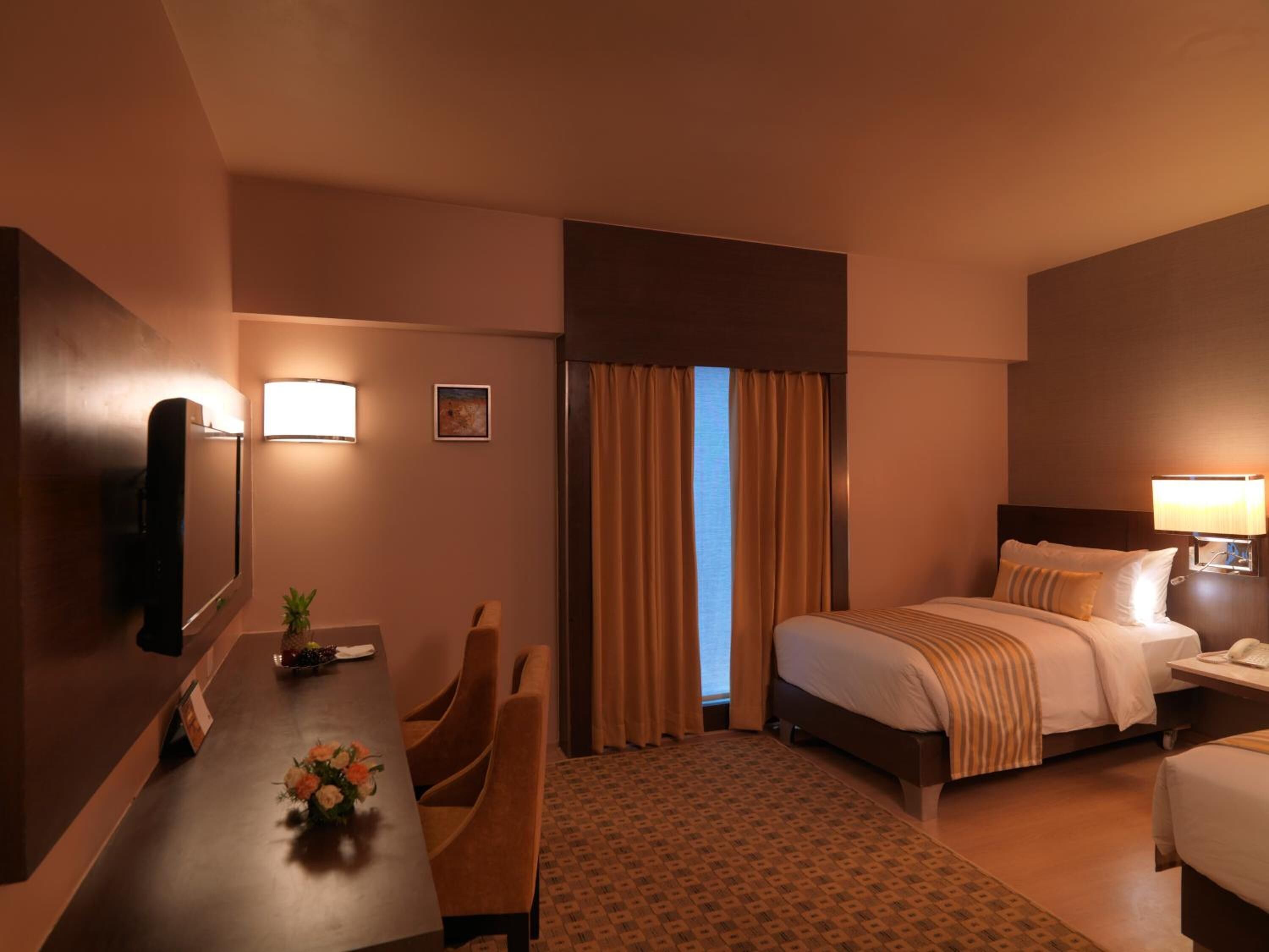 A hotel room with a bed, bedside table, lamp, TV, desk with chairs, and a window with curtains.