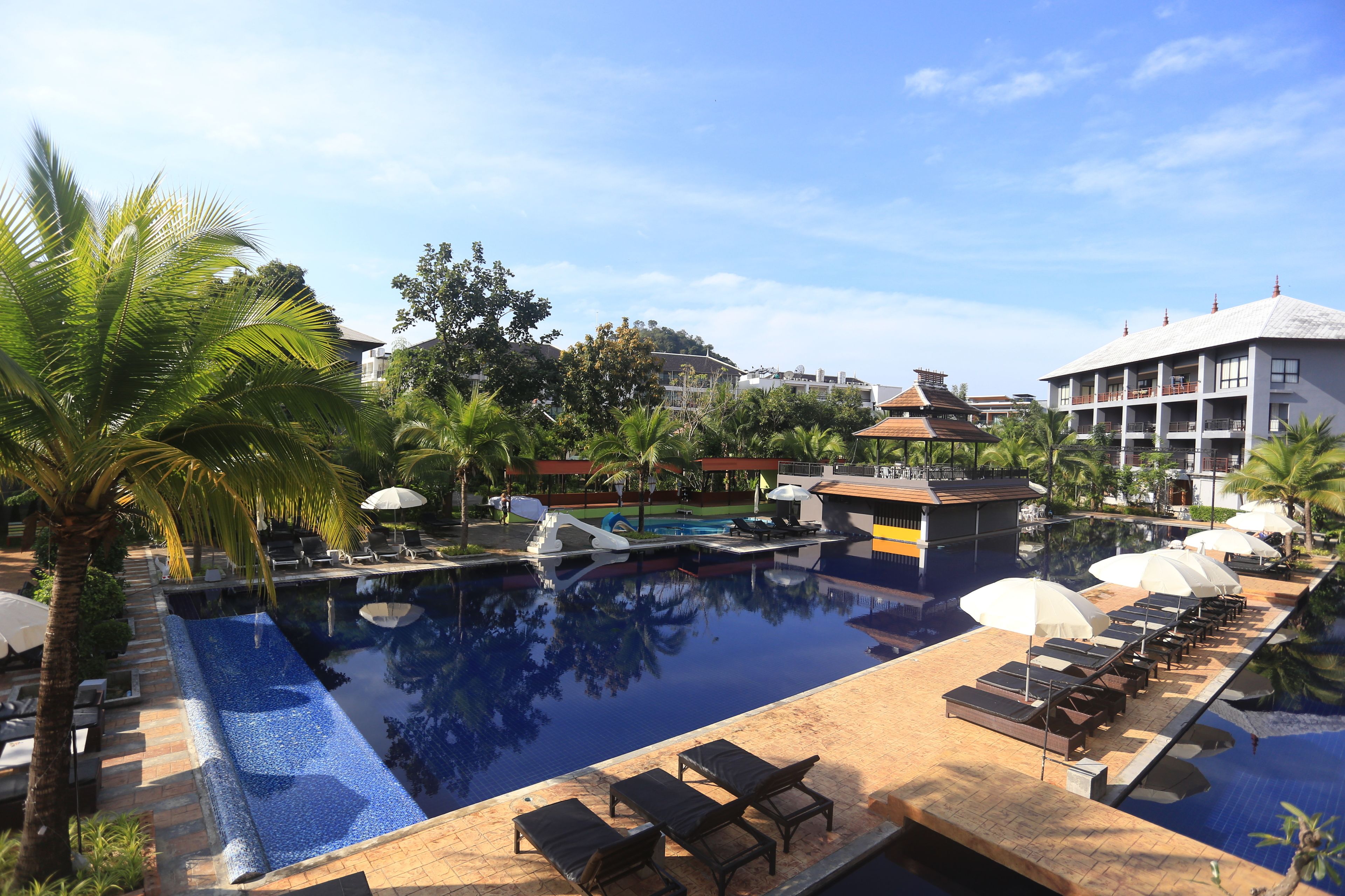 A resort pool area with lounge chairs and umbrellas, surrounded by palm trees and a building with a tiled roof.