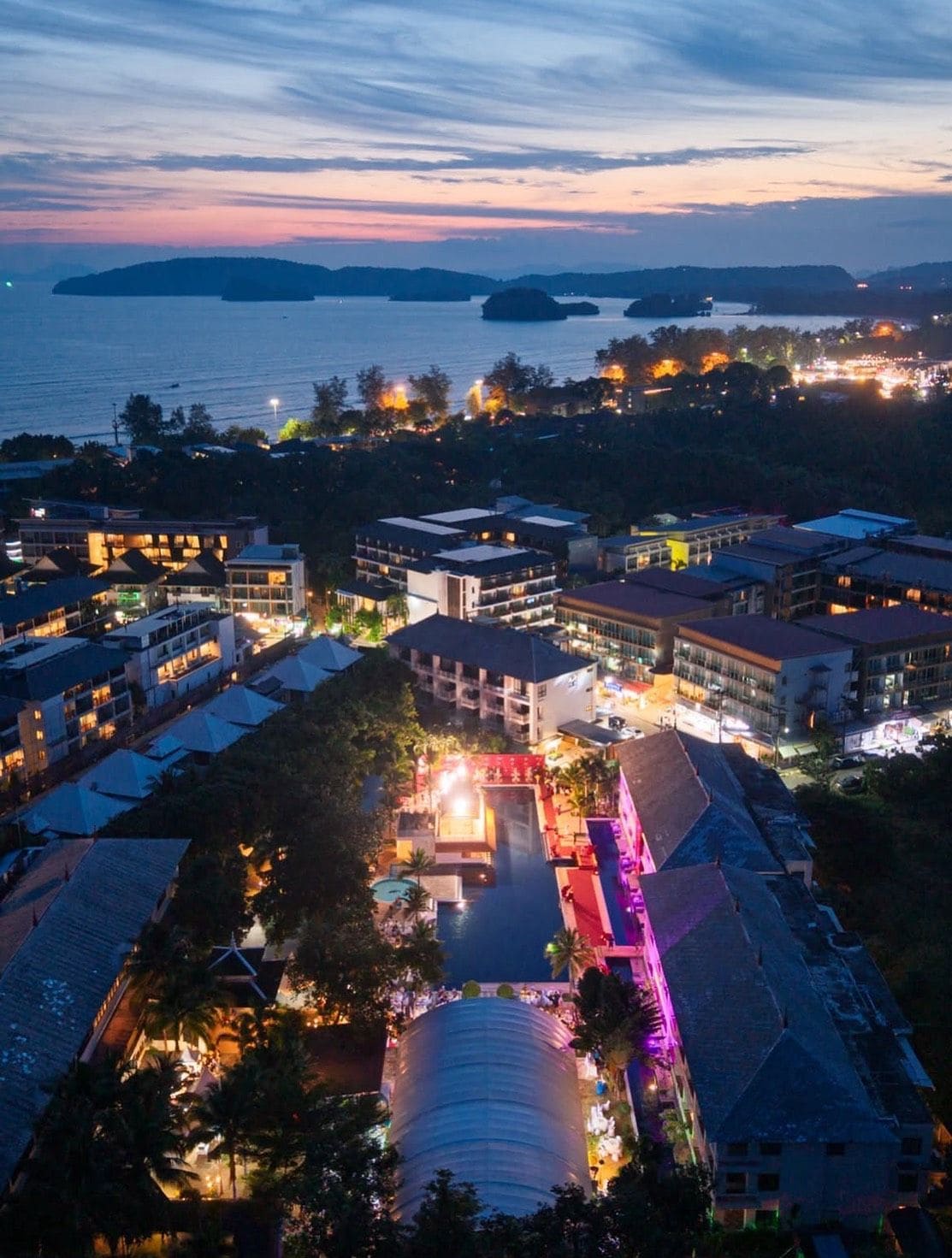 A resort with multiple buildings, a swimming pool, and a view of the sea at dusk.