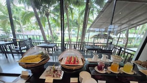 Un buffet de petit-déjeuner proposant des fruits tropicaux, des viennoiseries et diverses boissons, le tout servi dans une salle dotée de grandes fenêtres donnant sur des palmiers.