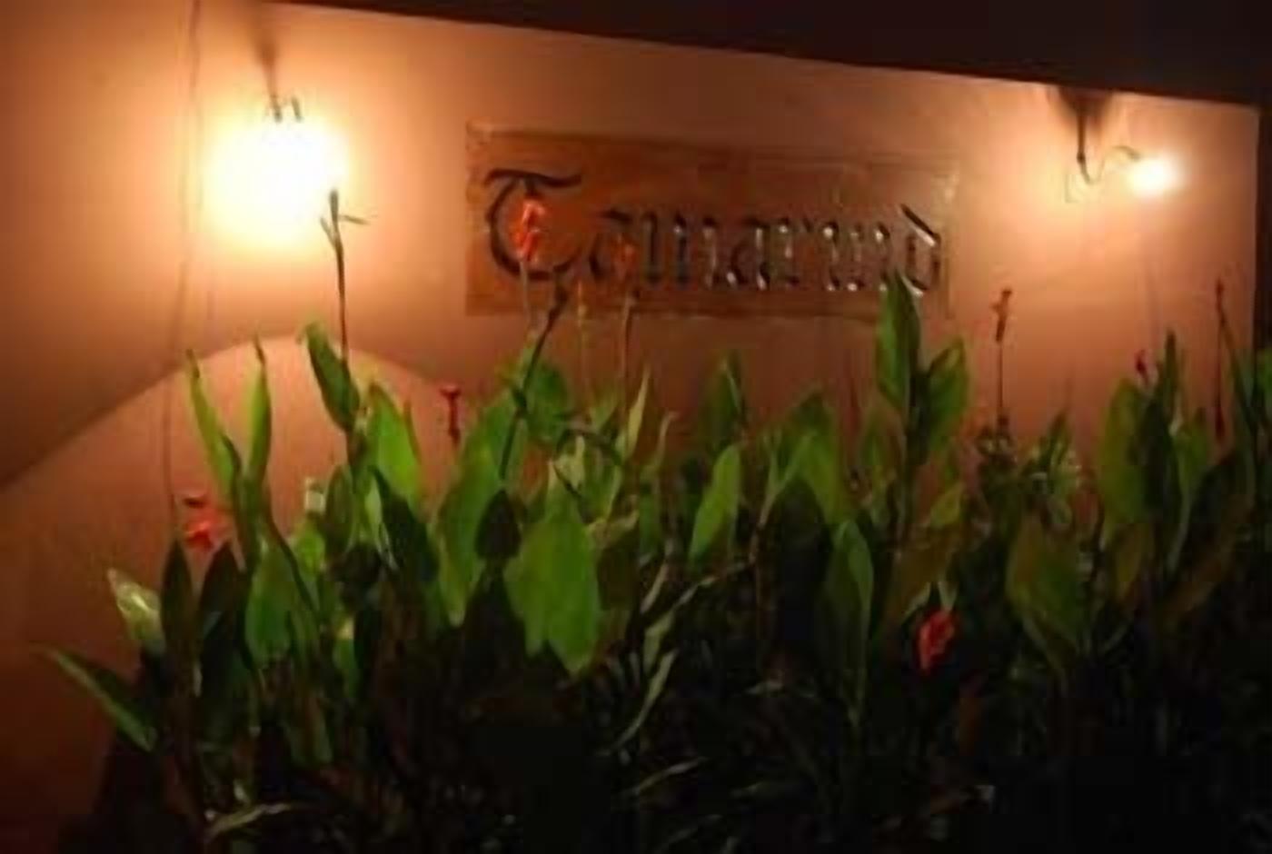 A sign with the name "The Garden Hotel" illuminated by wall-mounted lights, surrounded by lush green foliage.