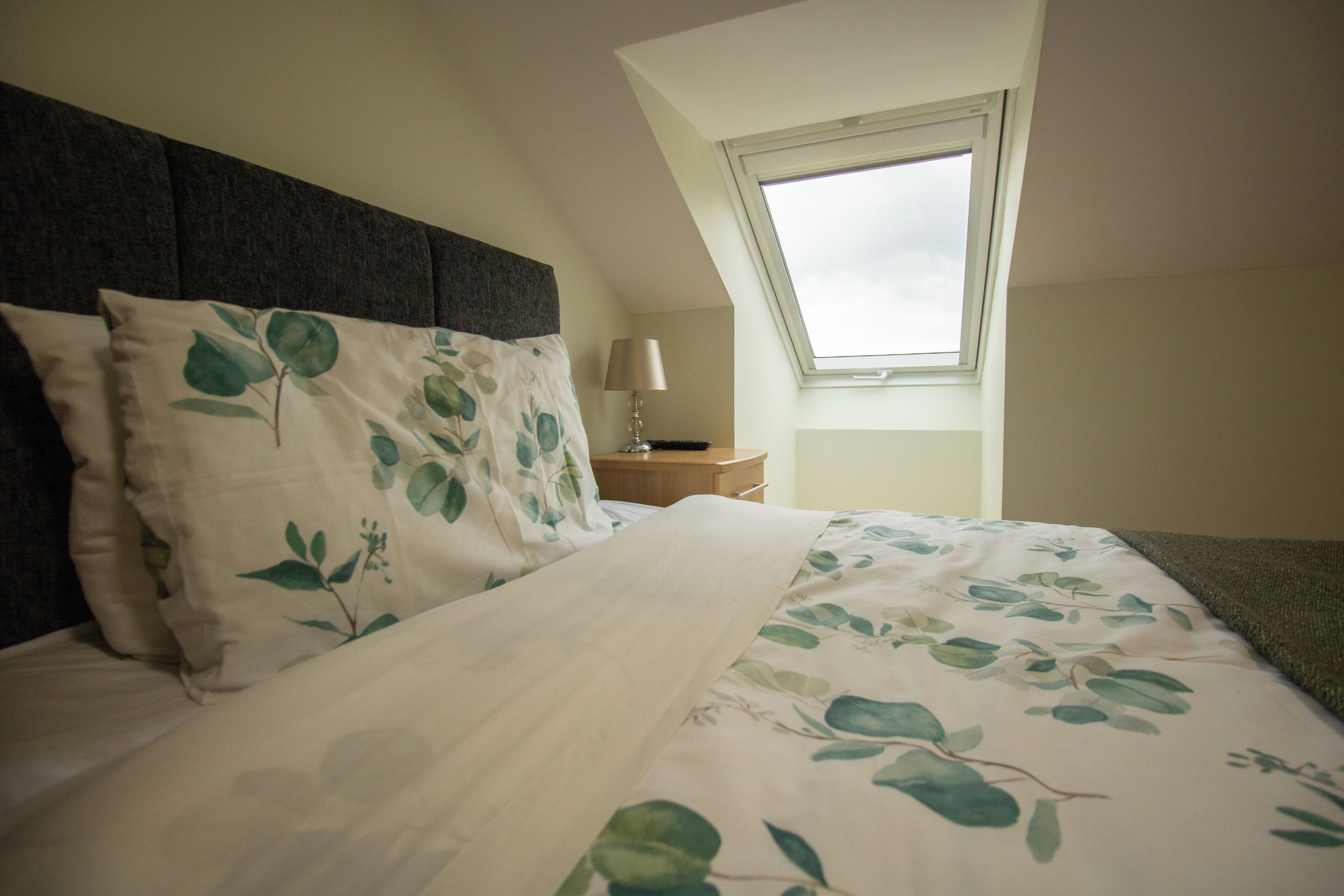 A bedroom with a bed, a nightstand, a lamp, and a skylight.