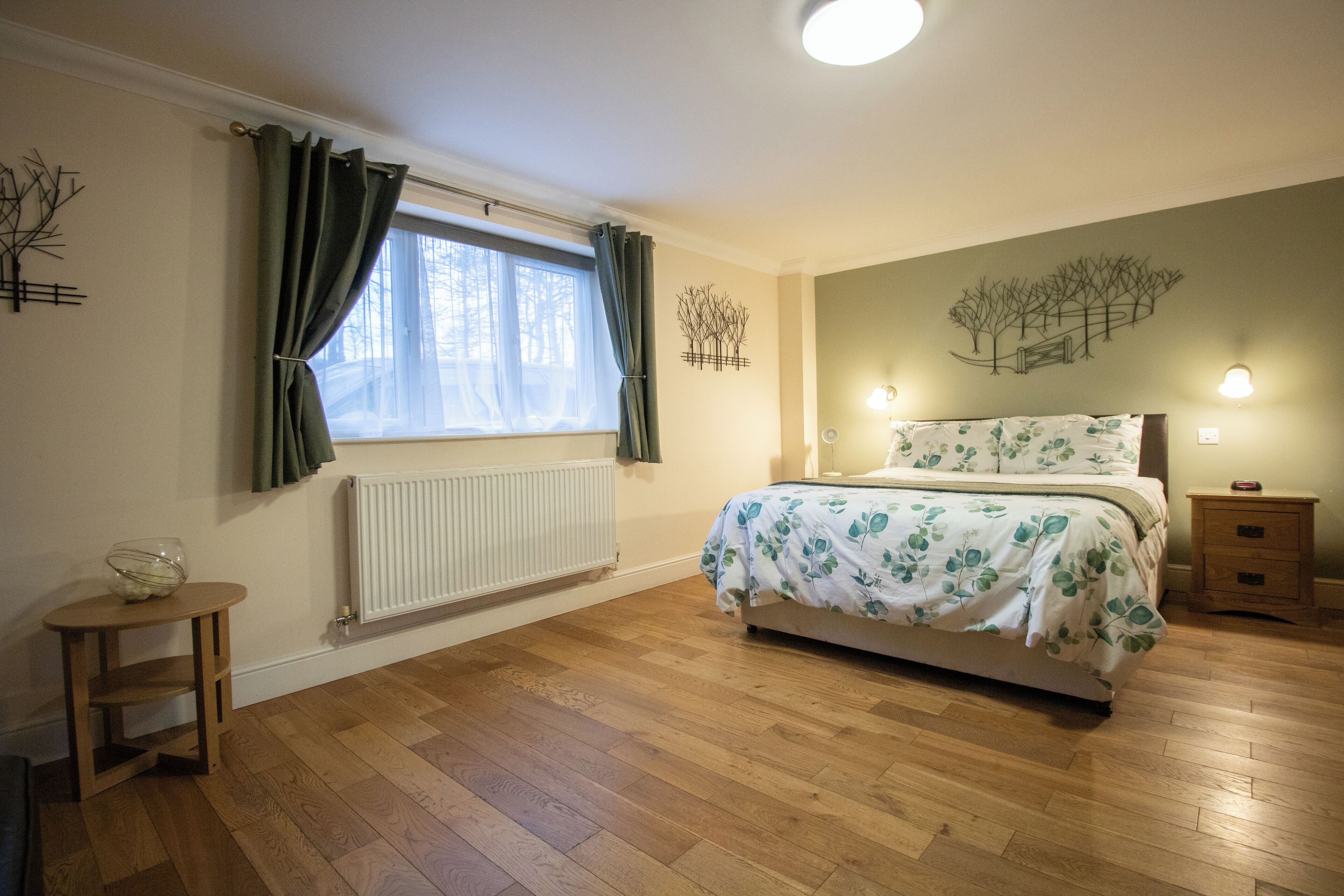 A bedroom with a wooden floor, a bed with a floral bedspread, a bedside table, a radiator, and a window with curtains.