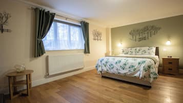 A bedroom with a wooden floor, a bed with a floral bedspread, a bedside table, a radiator, and a window with curtains.