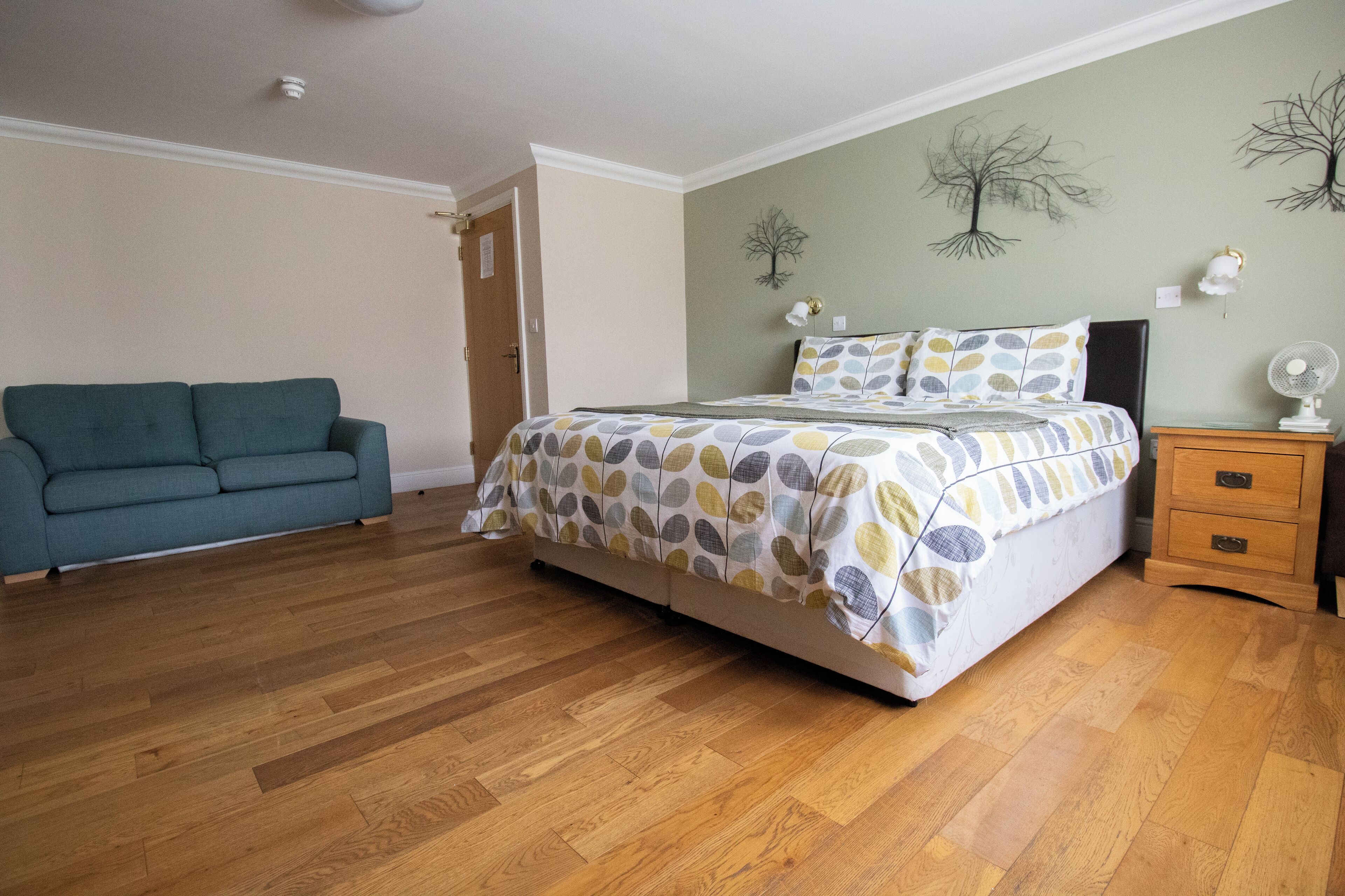Un dormitorio con cama, cómoda, mesita de noche, sofá y un muro decorado con motivos de árboles.