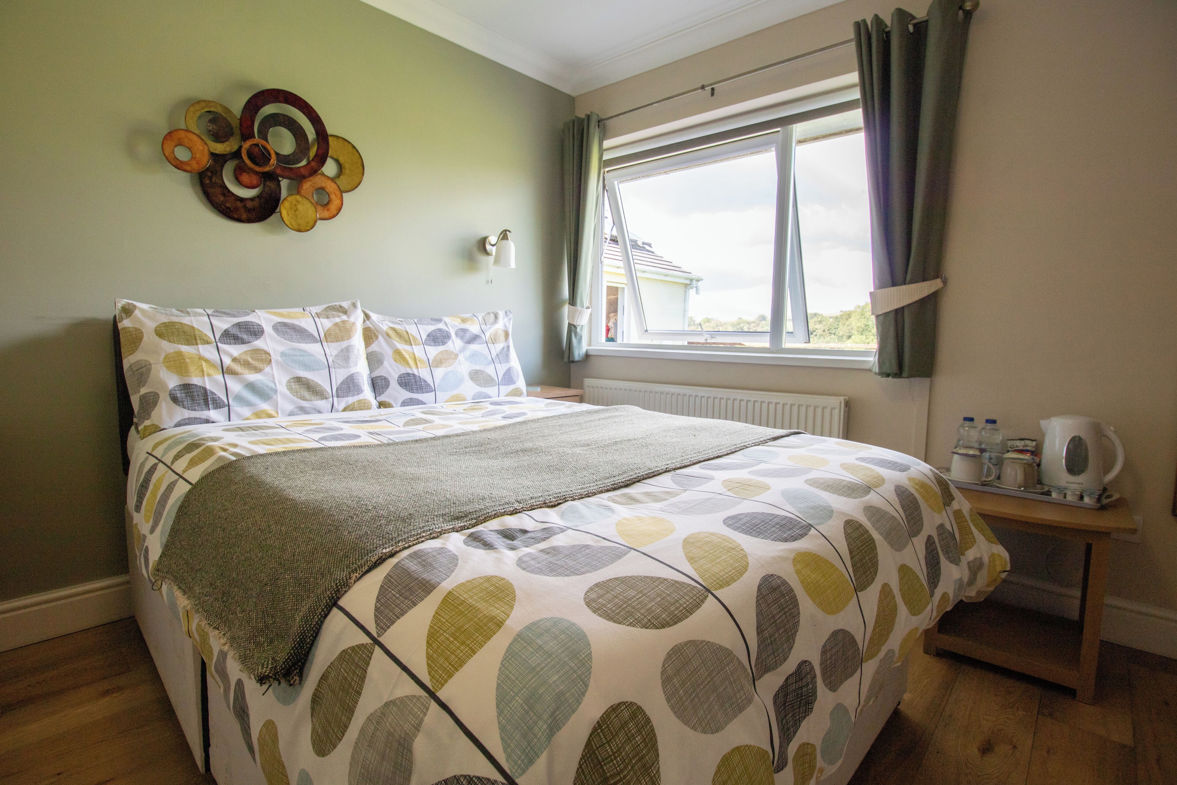 A bedroom with a bed, a window with curtains, a wall decoration, and a bedside table with a kettle.