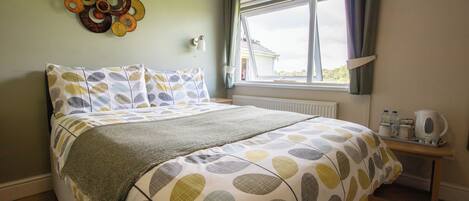 A bedroom with a bed, a window with curtains, a wall decoration, and a bedside table with a kettle.