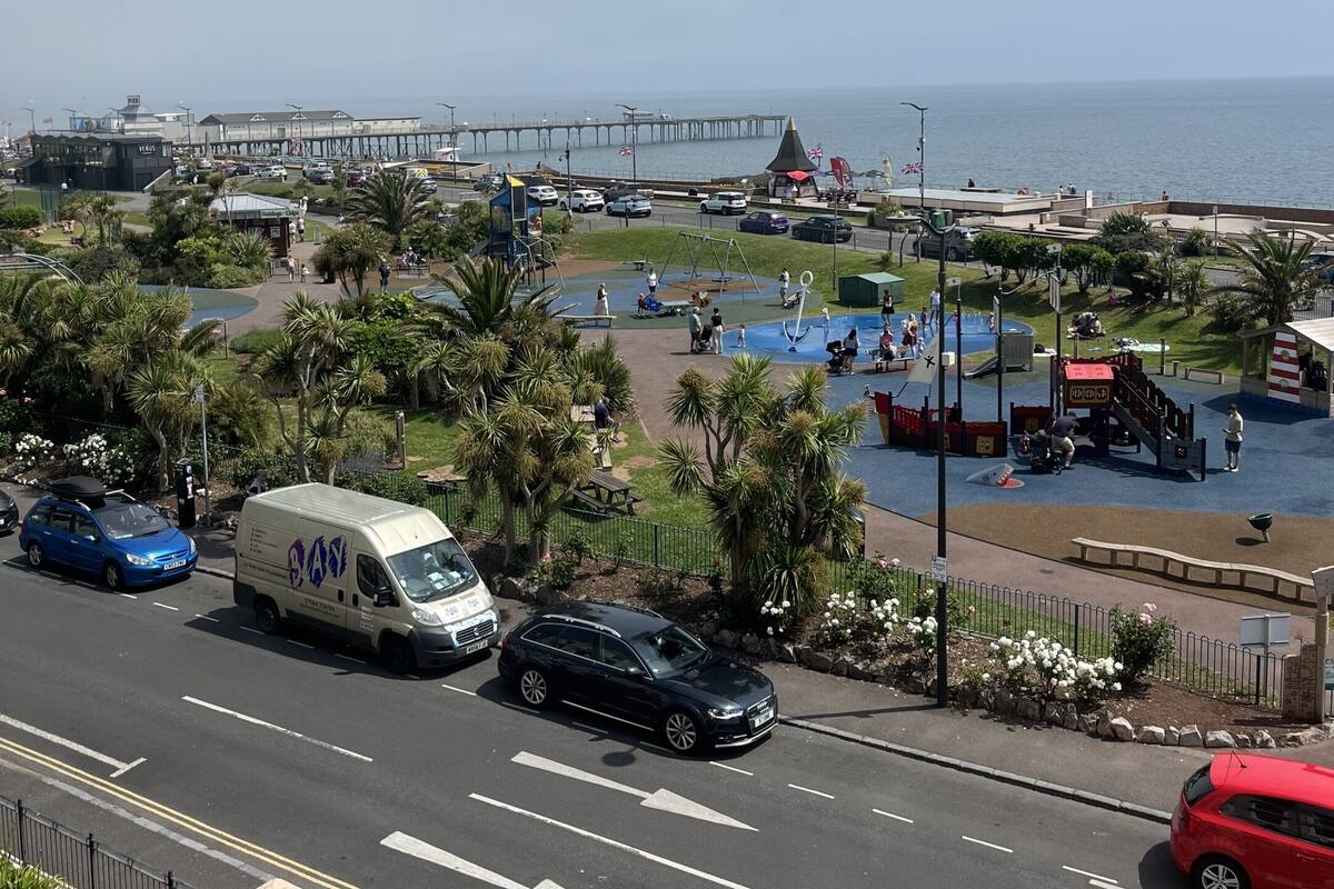 A coastal road with vehicles, a pedestrian area with playgrounds, and a pier in the background.