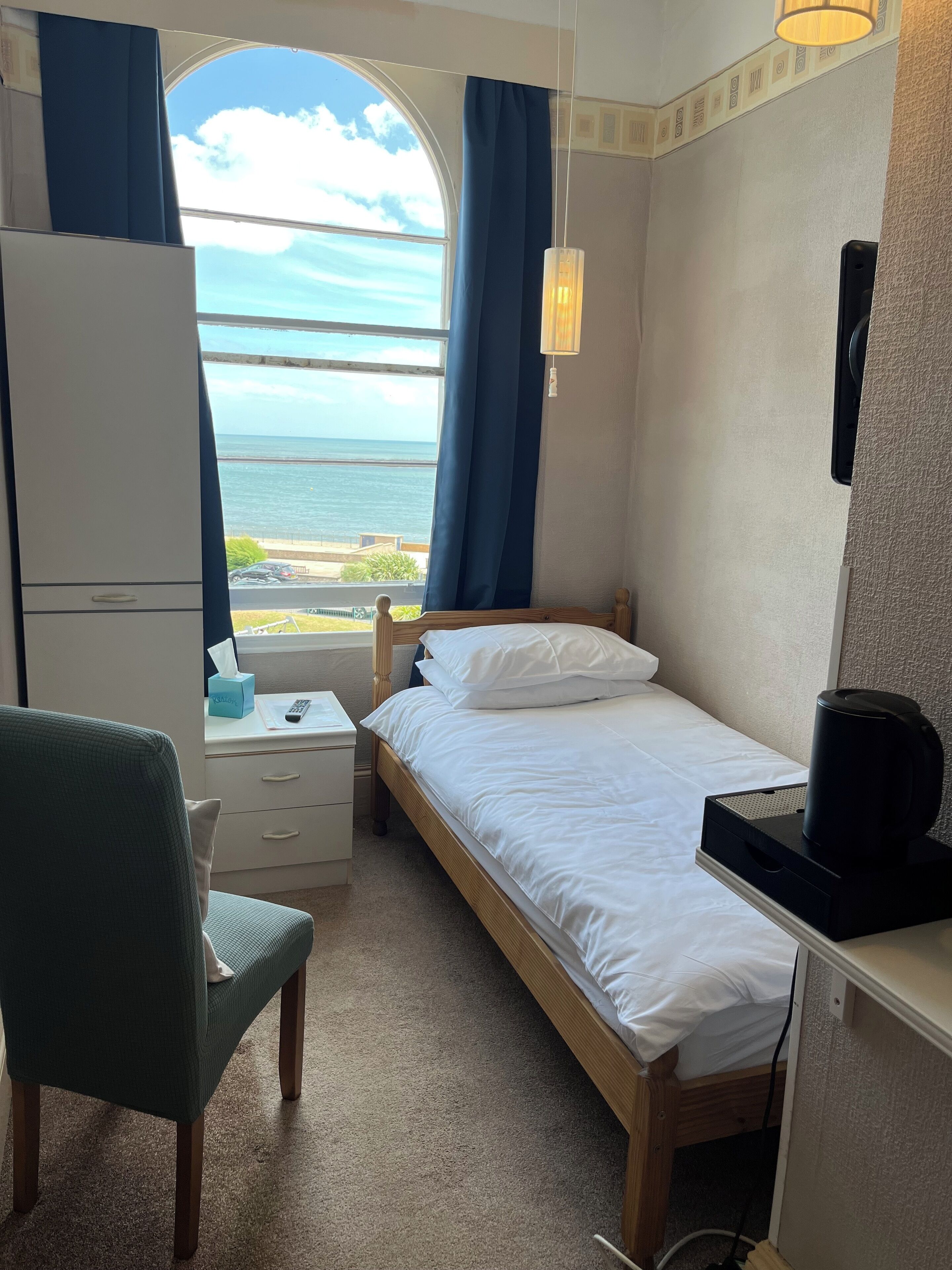 Ein kleines, ordentliches Schlafzimmer mit einem Einzelbett, einem Sessel, einem Schreibtisch und einem Fenster mit Meerblick.
