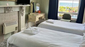 A hotel room with two beds, a vanity with a mirror, a television, and a view of the sea. - Lynton House (Teignmouth)
