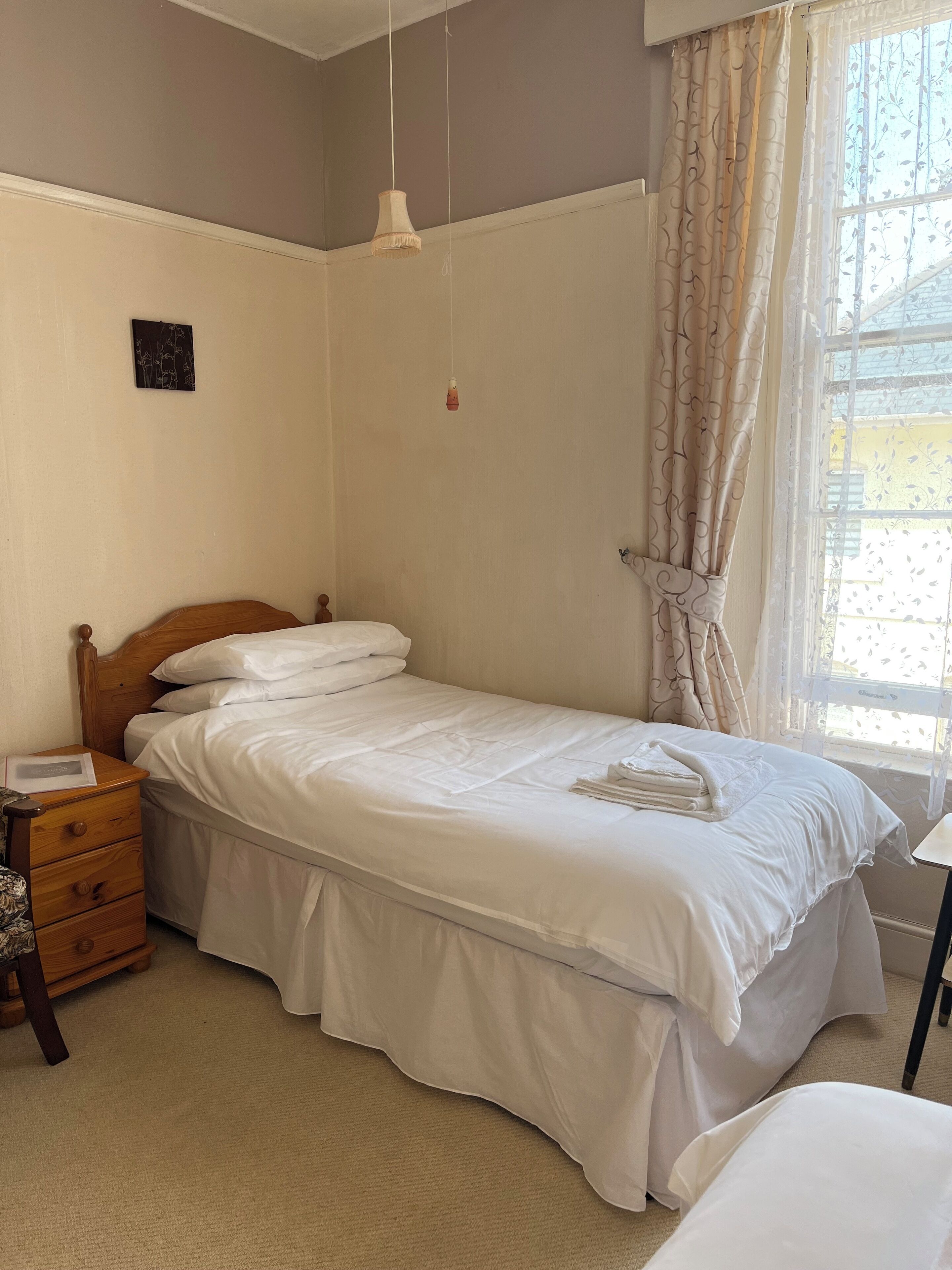 A neatly made bed with white linens, a wooden nightstand with a lamp, and a window with curtains.