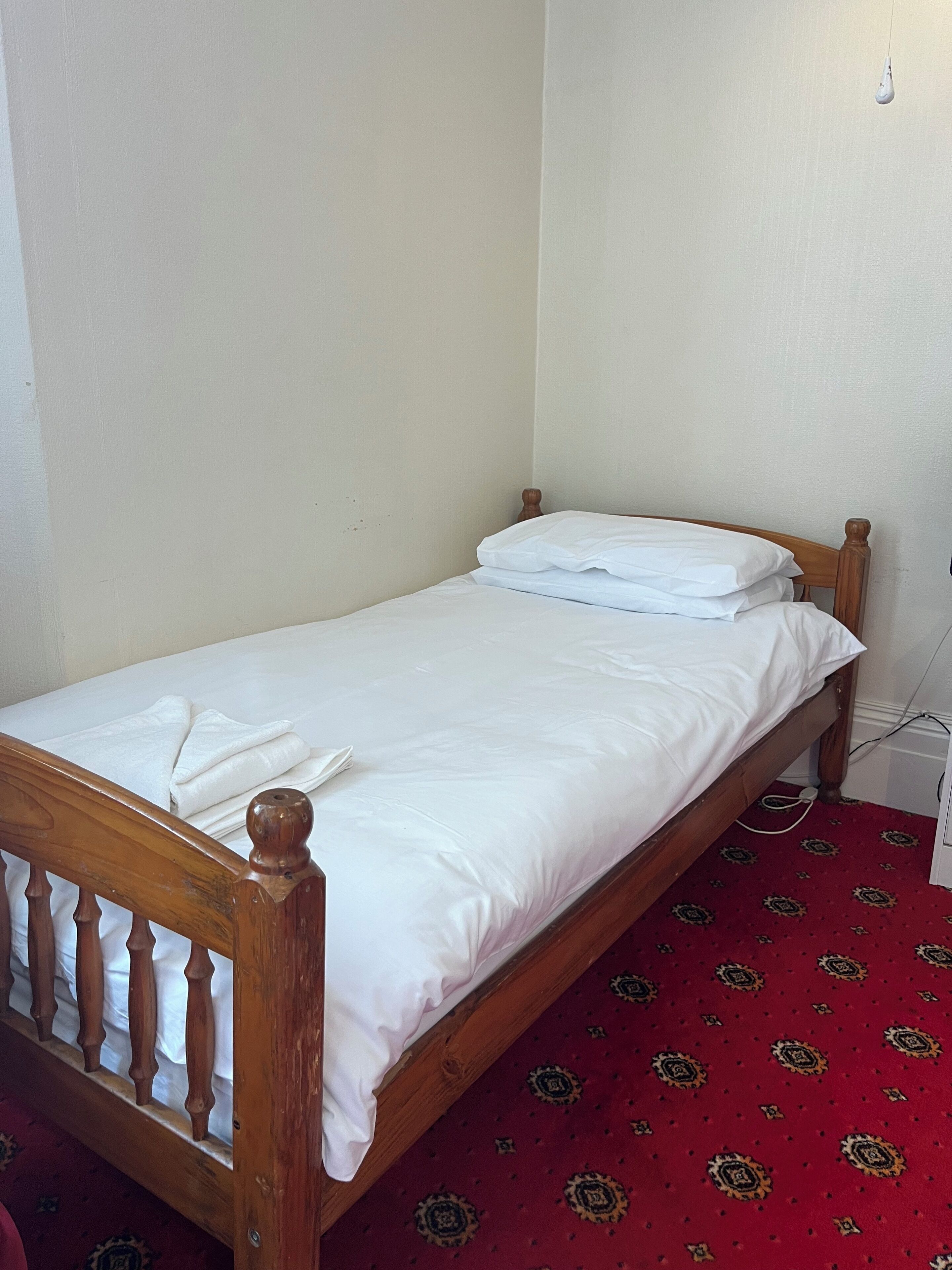 A small, single-bed room with a wooden headboard, white bedding, and a red patterned carpet.