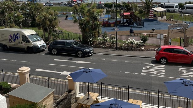 A coastal view with a road, parked cars, and a pedestrian area with benches and umbrellas. - Lynton House (Teignmouth)