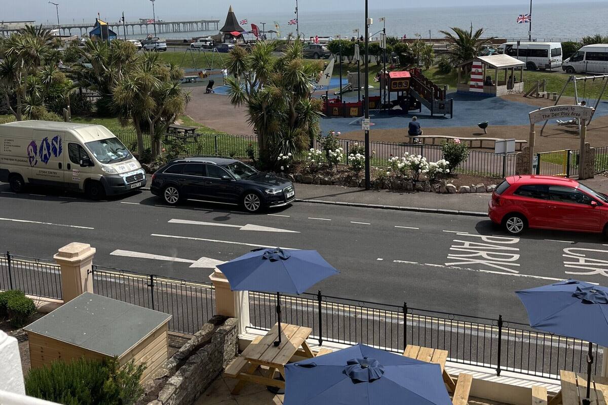 A coastal view with a road, parked cars, and a pedestrian area with benches and umbrellas.
