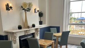 A room with a fireplace, a dining area with wooden tables and chairs, a large window with a view of the outside, and wall-mounted lighting fixtures. - Lynton House (Teignmouth)